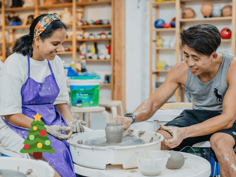 A woman in a purple apron smiles as she sits beside a pottery wheel with a clay pot on it alongside a male pottery tutor who is also smiling