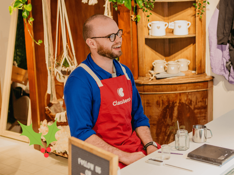 A man with glasses and a red ClassBento apron smiles widely as he sits next to a candle jar ready to fill with soy wax