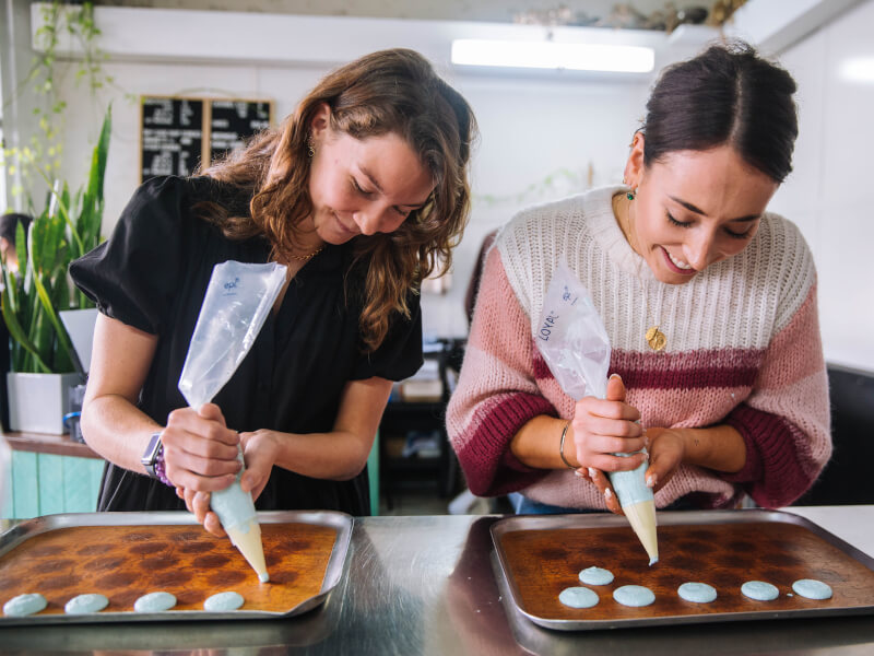 Two women are at a baking class, piping blue and pink macarons onto baking trays