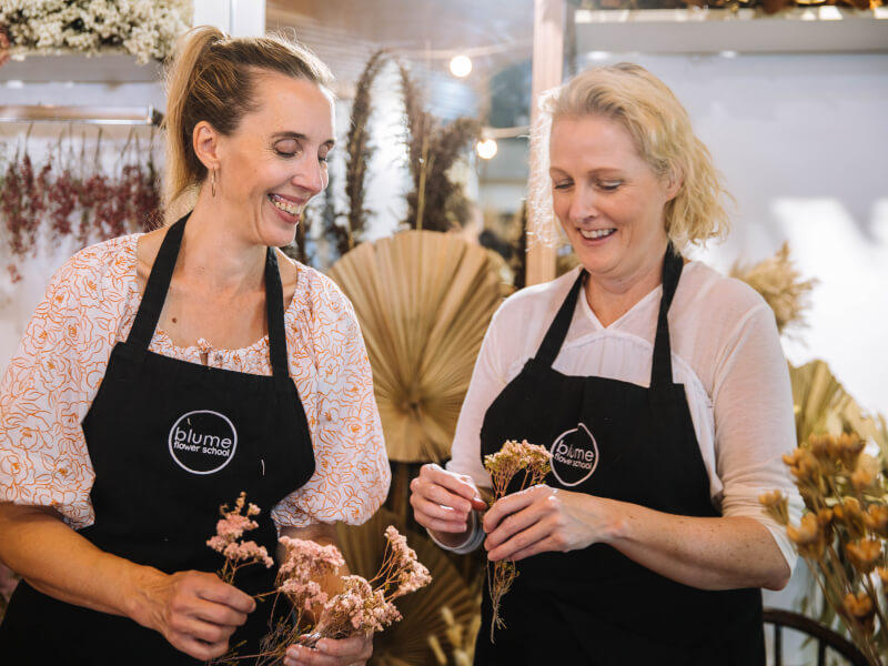 Two women wearing aprons holding flowers and smiling.