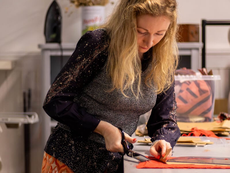 A woman stands at a large table, cutting orange fabric into pieces