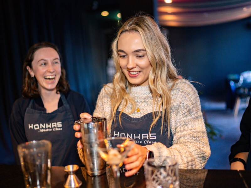 A smiling woman is making cocktails in a cocktail shaker, another woman is standing behind her laughing