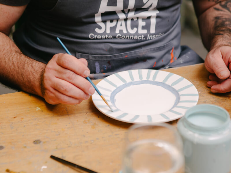 A close up of a person painting a blue stripe design on a white ceramic plate
