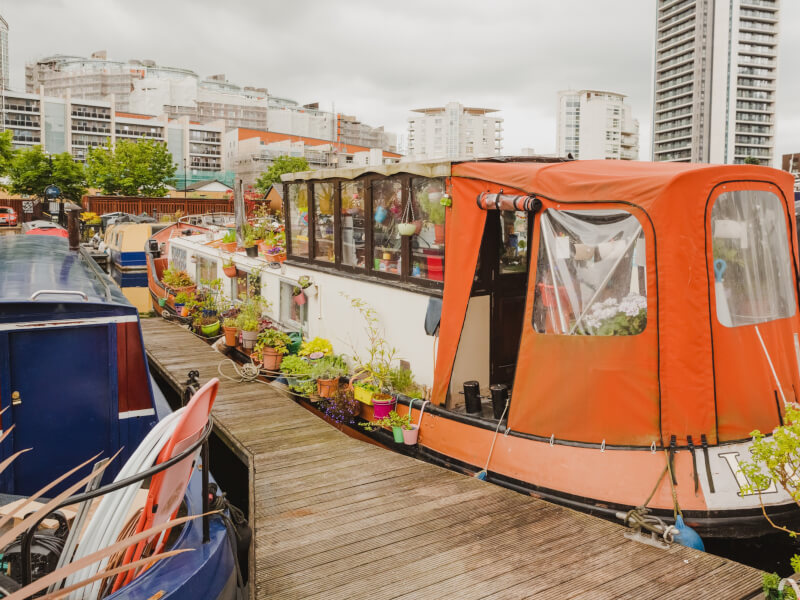 A houseboat covered in plants is docked in London, with buildings in the background