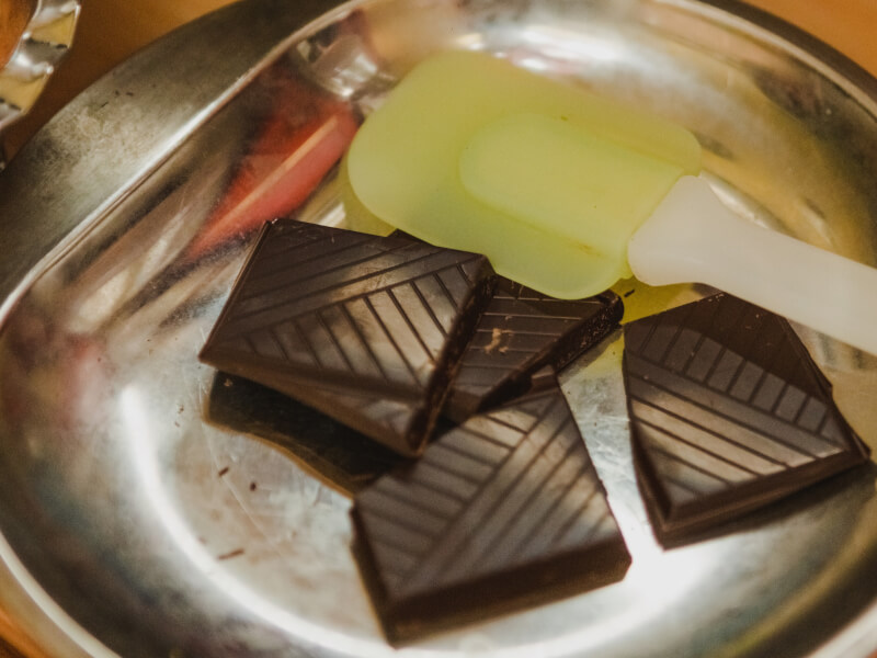 Close up of a metal bowl and green rubber spatula stirring squares of dark chocolate