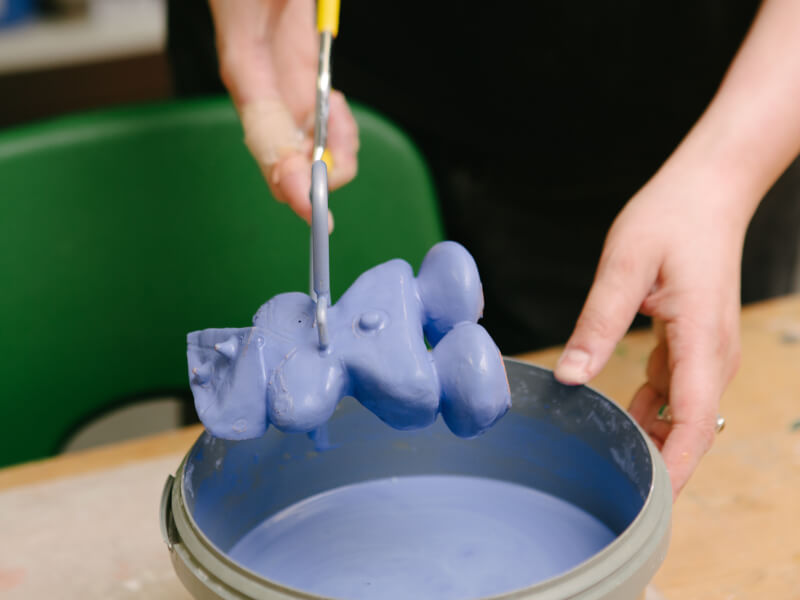 Person using tongs to remove a ceramic figure from a vat of lilac paint