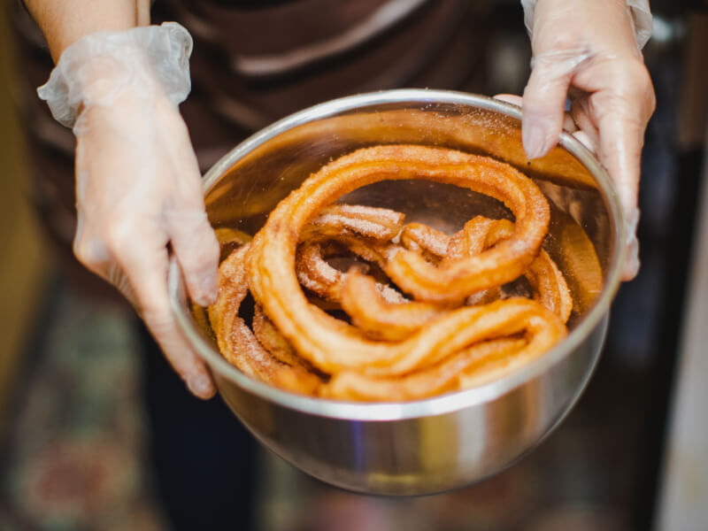 Close up of hands in food prep gloves holding a stainless steel bowl containing crispy churros sprinkled with sugar