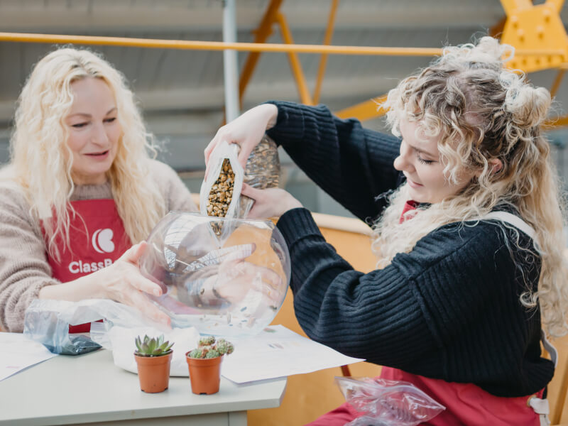 Two women making a terrarium together whilst wearing pink aprons. 