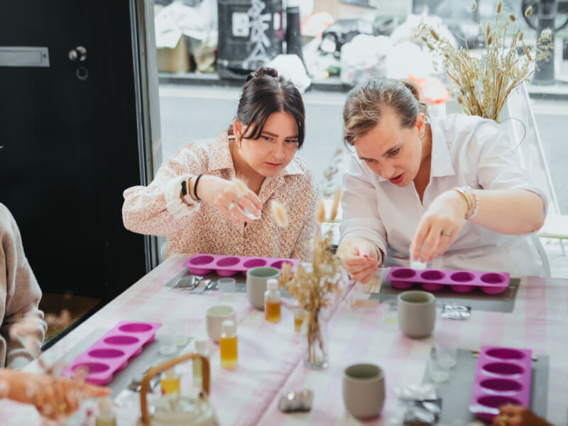 Two women at a soap making class by Yougi sit at a table measuring liquid