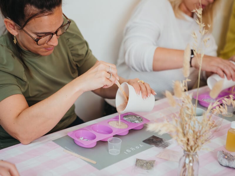 A woman in a green tshirt is pouring handmade soap into a pink silicon mould