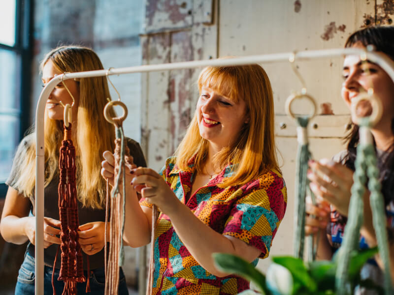 Three women standing at a clothes rack which is holding the beginnings of macrame plant hangers 