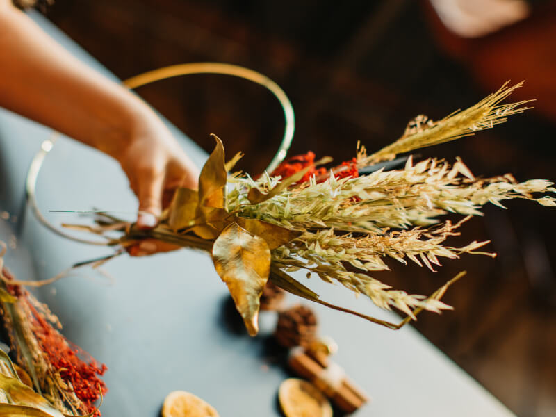 Dried flower selection of muted shades of green and brown being held by a hand in the background next to a golden ring hoop