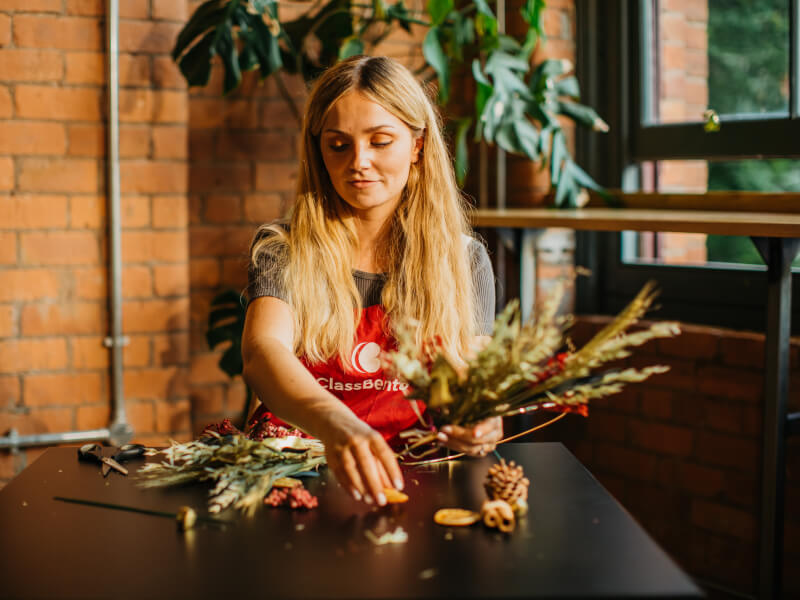 A woman sits at a table making a festive wreath with a selection of greenery and berries