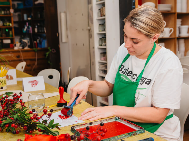 A woman sits at a table in a green apron, inking a lino block to print Christmas cards
