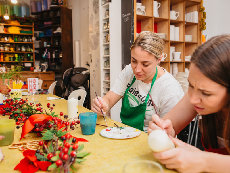 Two women sit at a table, painting white pillar candles with small colourful motifs