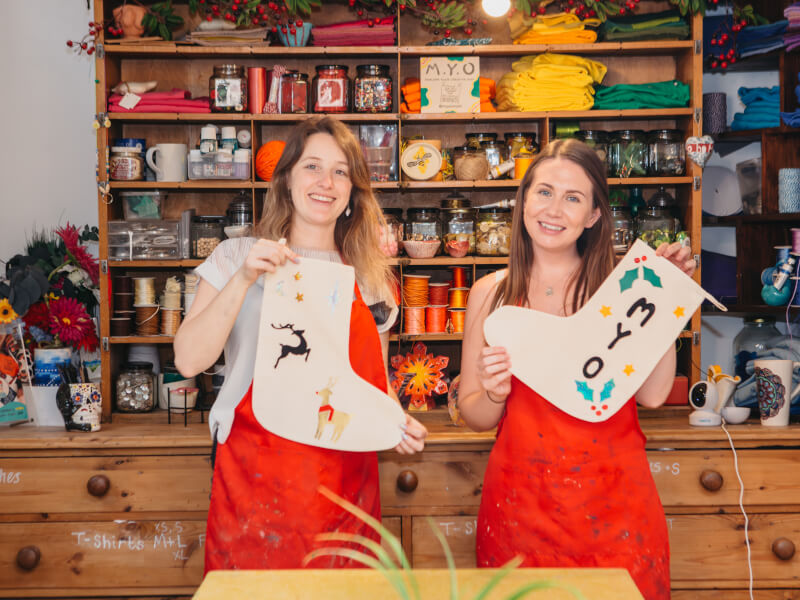 Two people are holding handmade Christmas stockings in a craft studio