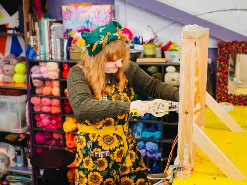 A person in colourful clothing using a tufting gun on a stretched canvas
