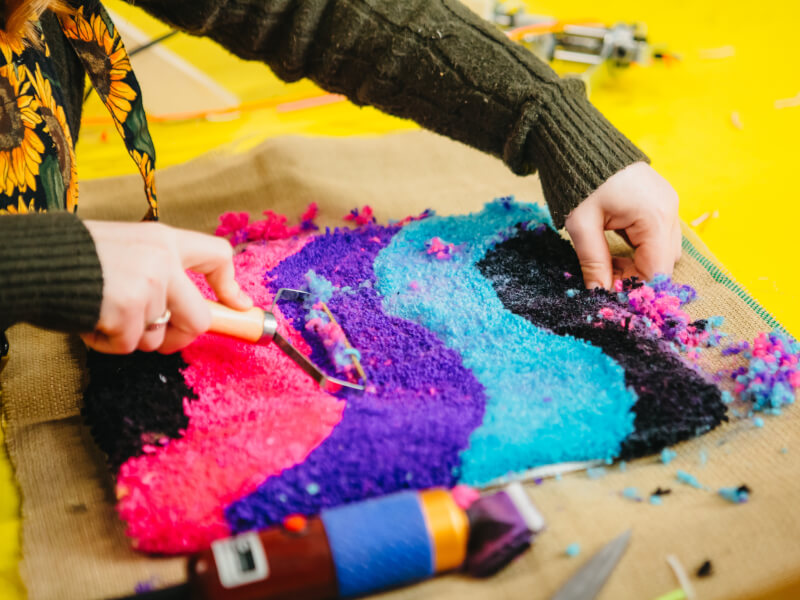 A close up of two hands trimming a freshly hand tufted rug, made of multi-coloured yarn