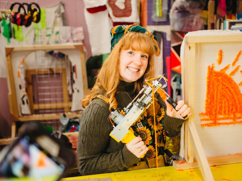 A woman holding a tufting gun with their half made rug in a tufting class.