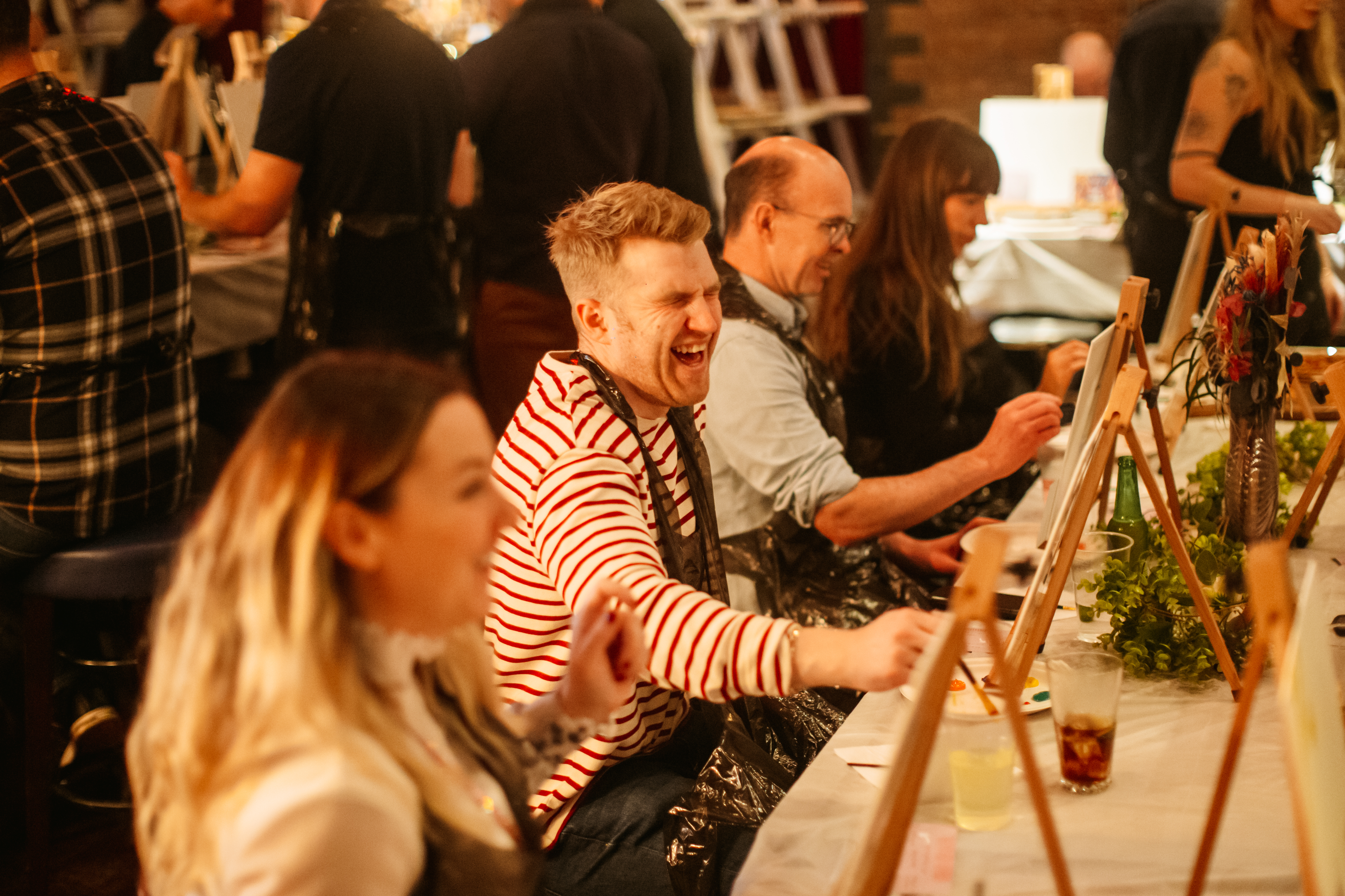 A man is sitting at an easel at a paint and sip class, laughing 