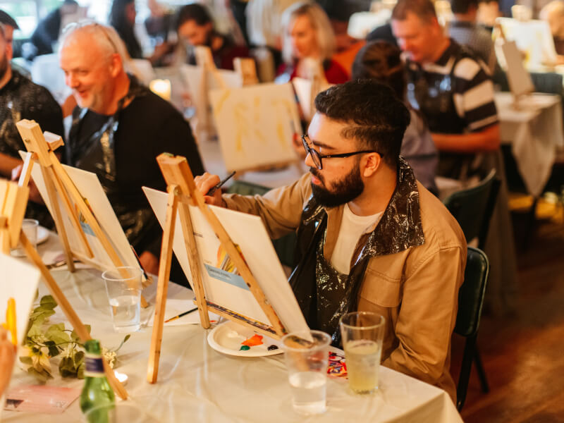A man adds paint to a canvas at a paint and sip event.