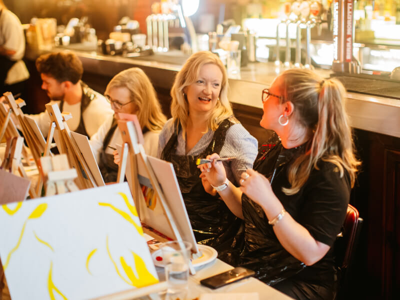 A group of laughing women are sitting at easels at a painting class