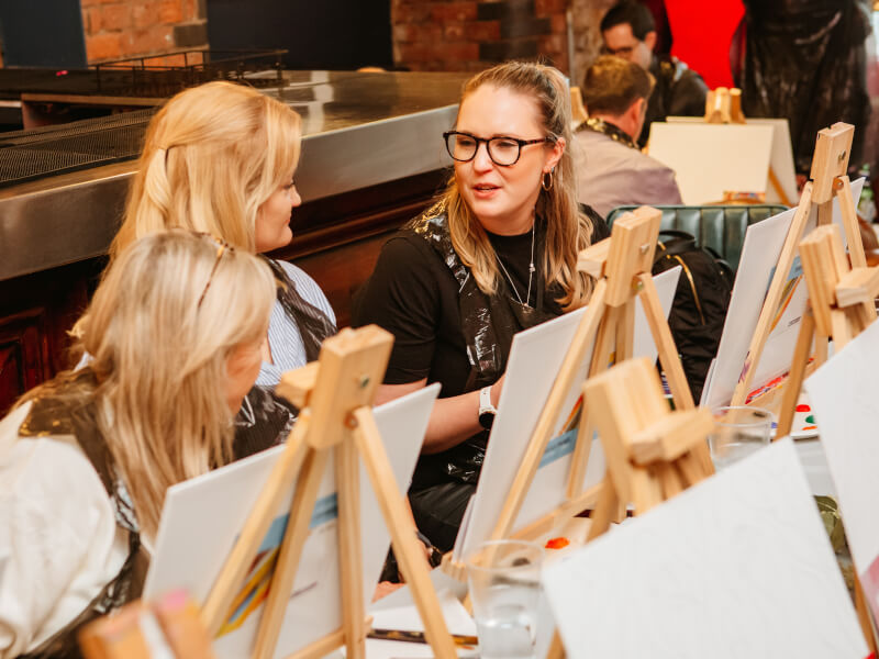 Three women sat behind easels at a paint and sip class.