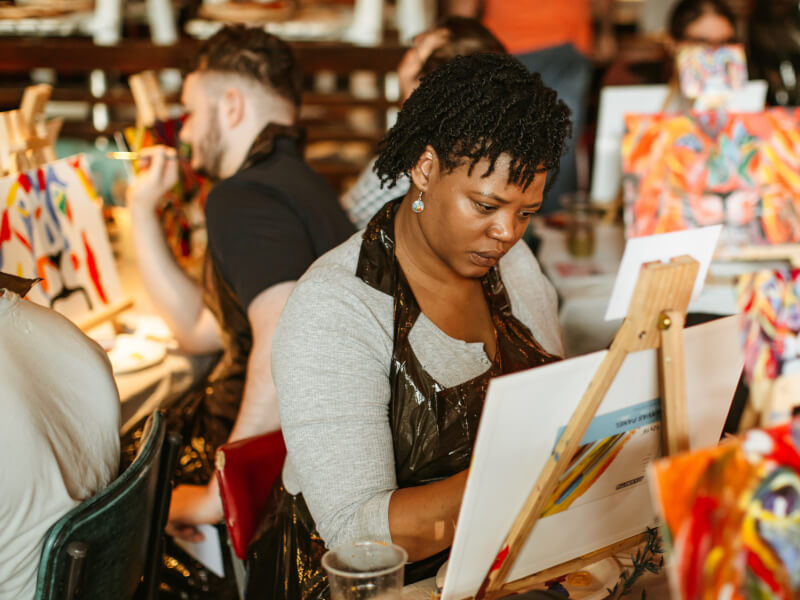 A woman concentrates as she sits and paints at an easel