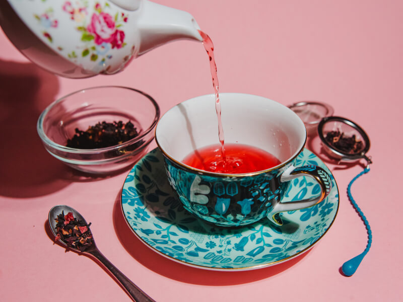 A tea pot pouring tea into a tea cup on a pink background.