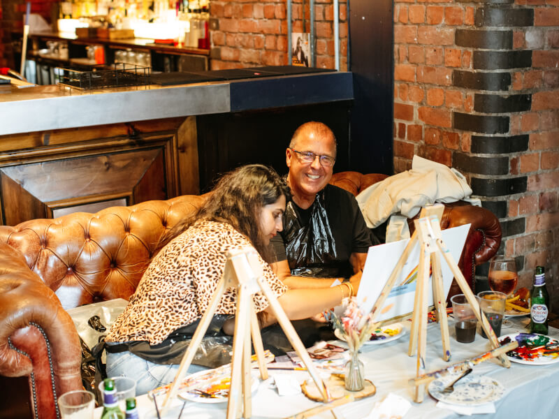 A man smiling whilst teacher helps him at an art class. 