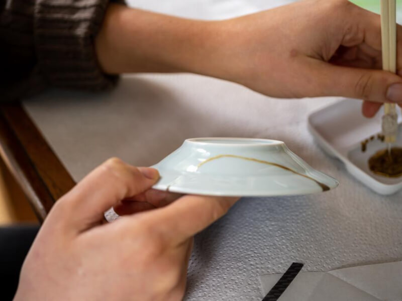 CLose up of hands holdig a white porcelain dish with a vein of gold kintsugi