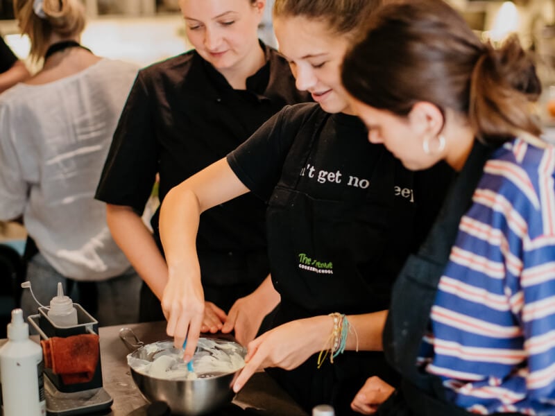 One women in a cooking class stirs something in a bowl whilst two people watch.