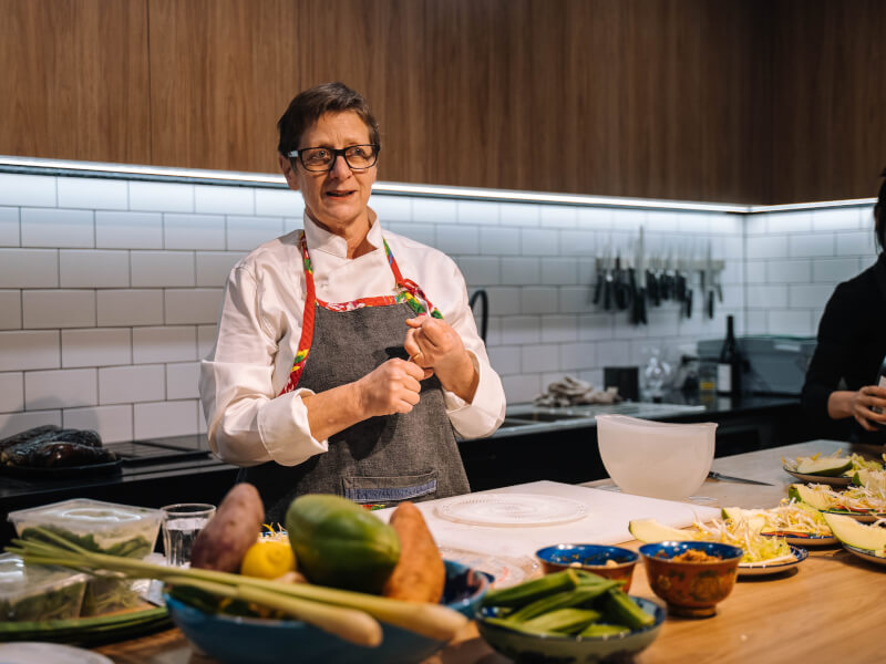A woman wearing an apron stood behind a kitchen bench filled with ingredients.