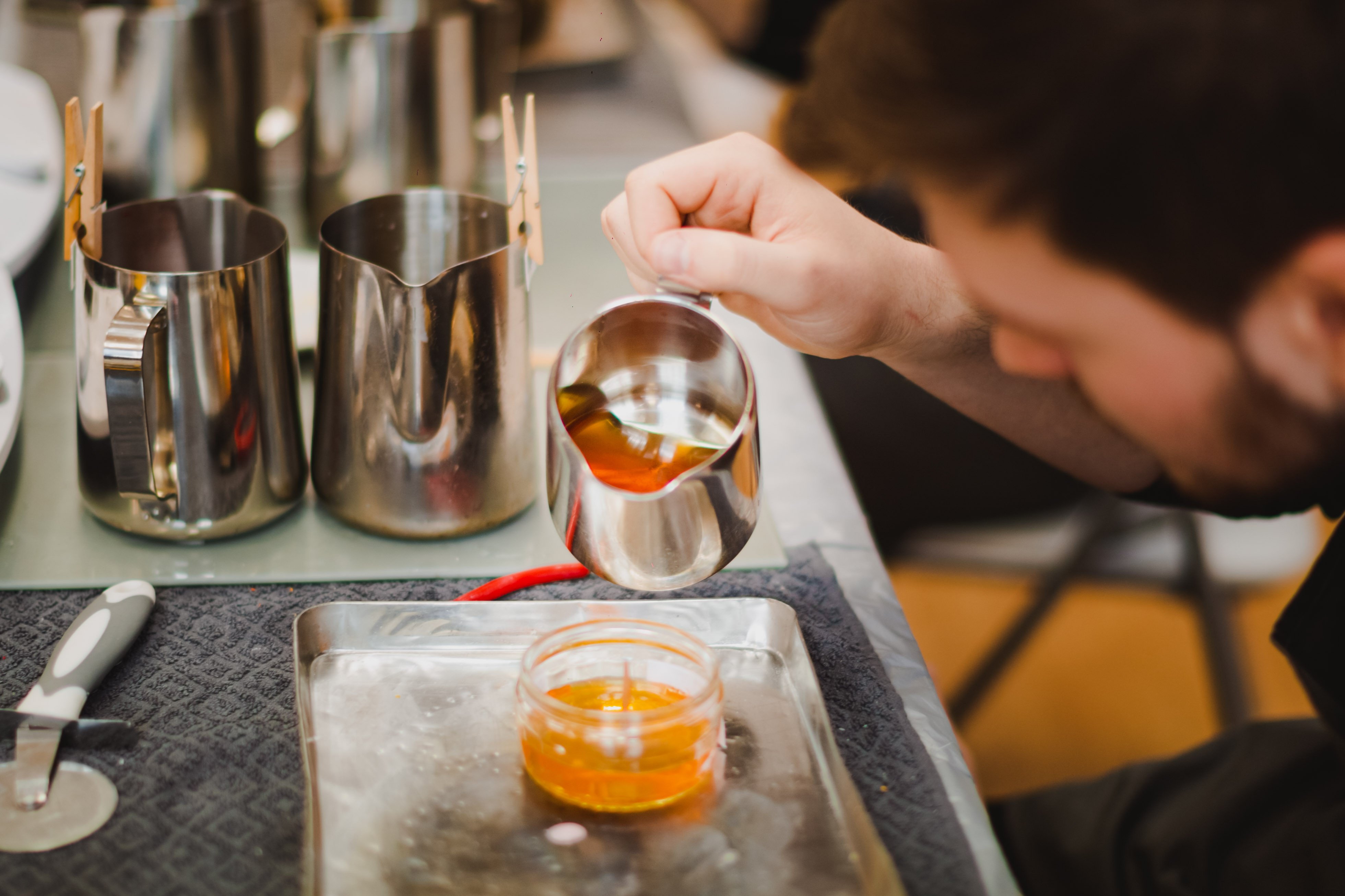 Close up of a man with a beard pouring wax from a metal jug into a container