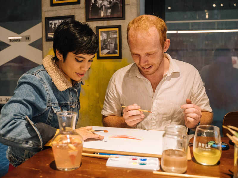 A man and woman sit at a table painting a picture together, with drinks nearby