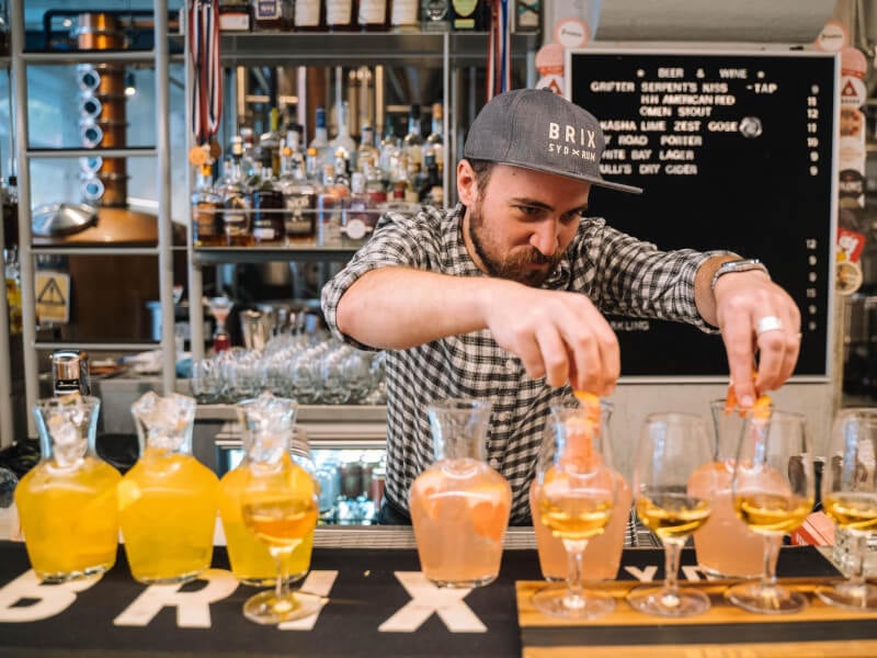 A man in a sports cap arranging garnishes on a row of orange cocktails