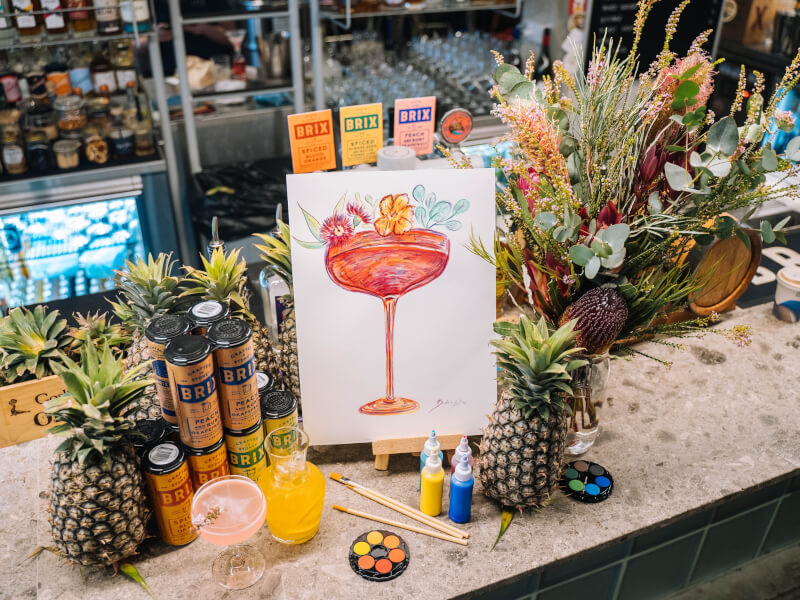 A painting of a cocktail propped up on a bar next to drinks and paint.