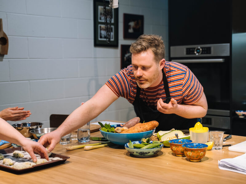 A man stretching across a table to reach some ingredients at a cooking class.