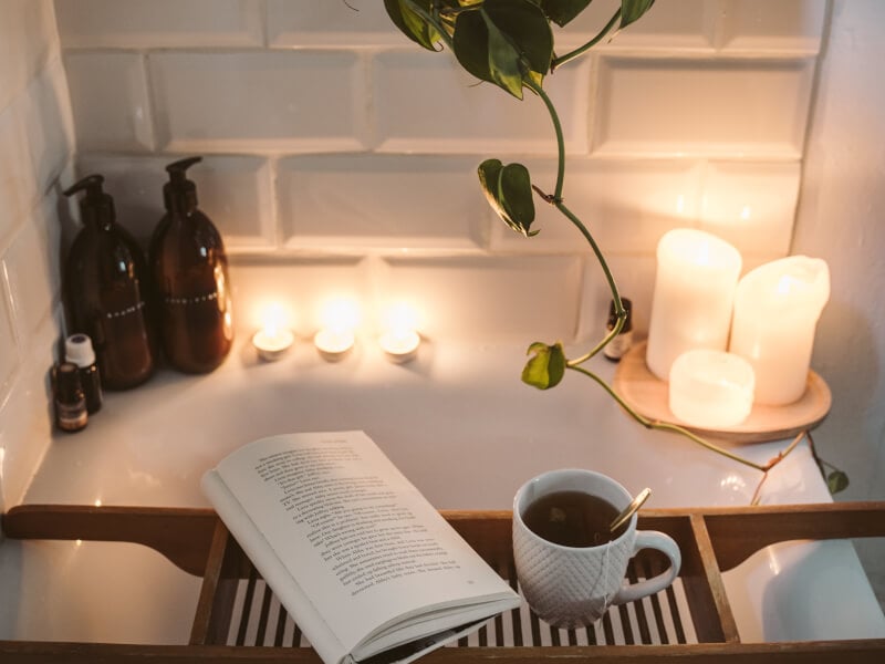 A bath with a book and tea resting on a bath caddy, surrounded by tall white candles