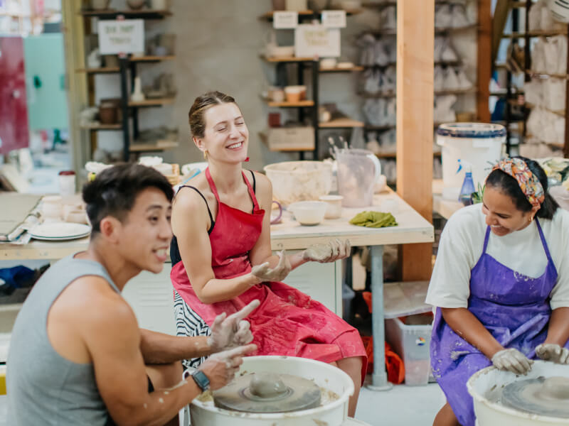 Three people laughing at their clay-covered hands as they sit at pottery wheels with a lump of clay