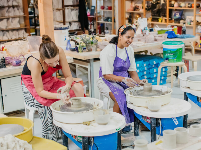 Two women are using pottery wheels side by side to make small vases