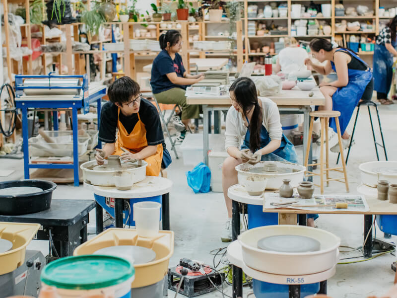 Two people looking at each other whilst both making pottery on a wheel. 