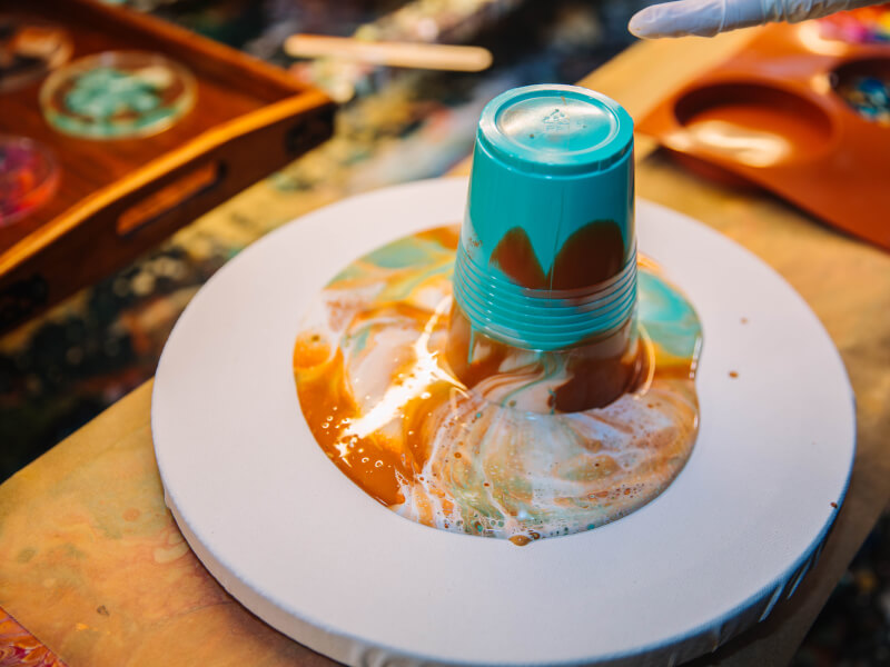 A cup pouring acrylic paints onto a canvas