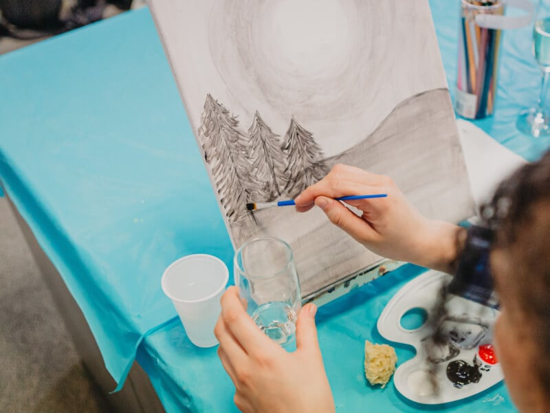 A person paints a forest scene at a drink and paint class