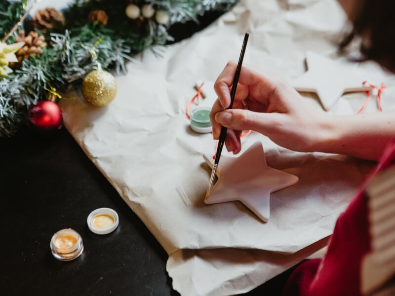A close up of a hand holding a paintbrush covered in yellow paint, painting a clay star
