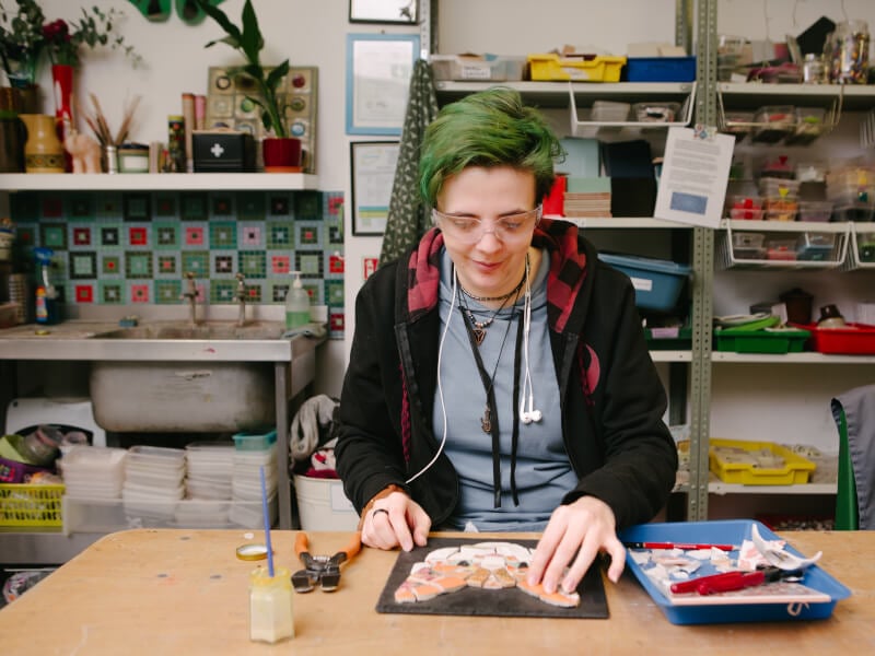 A person is sitting at a table creating a mosaic design of a cat's face using coloured tiles