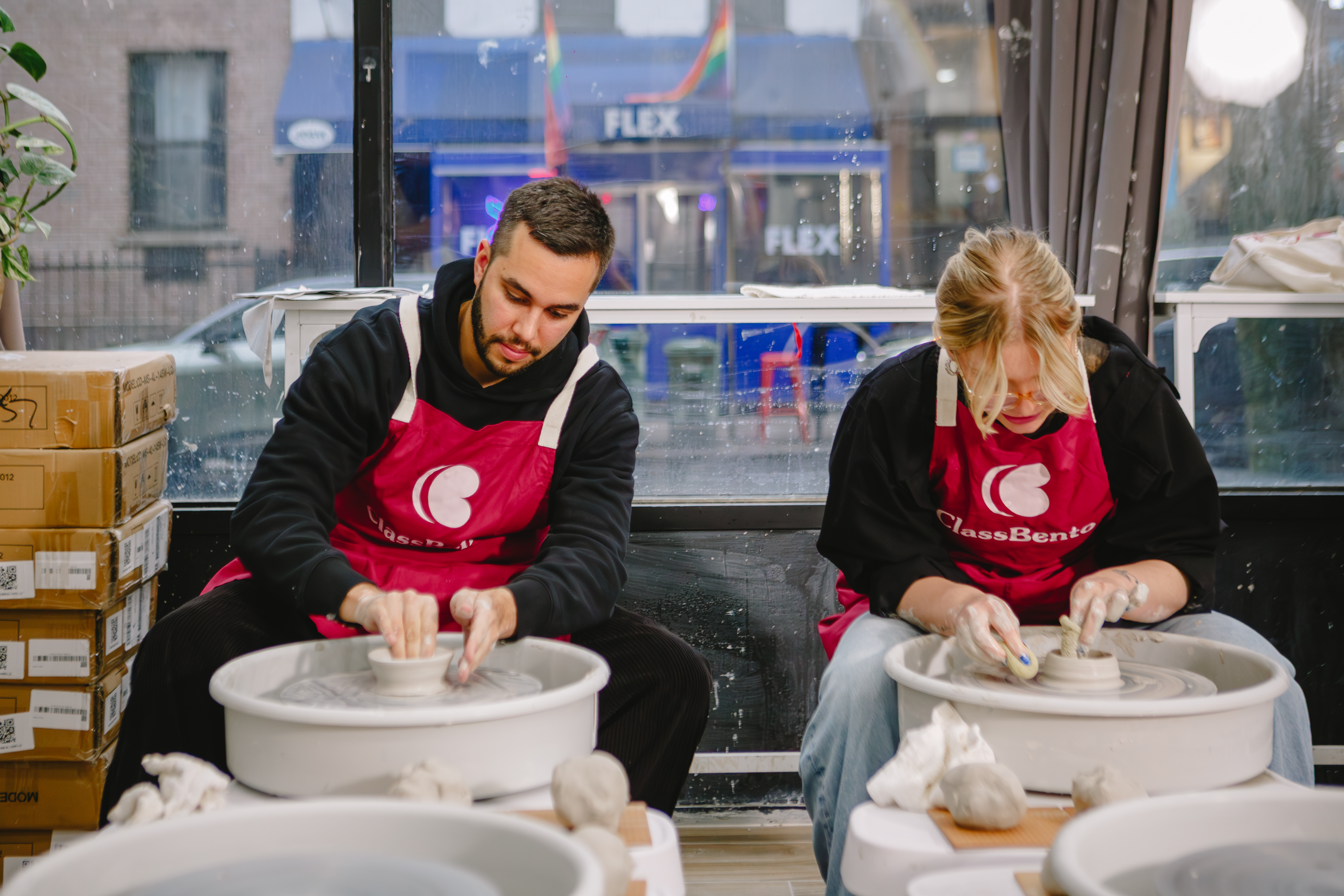 Two people are sitting at pottery wheels, making small pots from clay