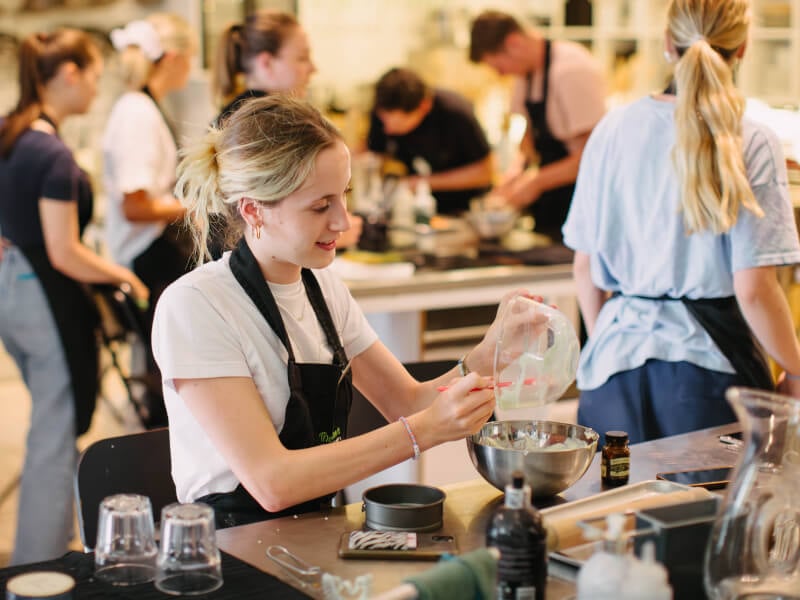 A woman sits at a table at a cooking class, pouring a mixture from one bowl to another