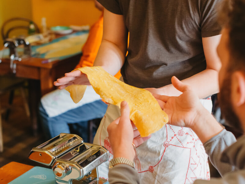 Two men hold a sheet of handmade pasta from their pasta making machine.