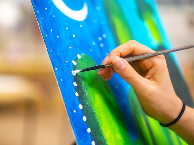 A close up of a hand adding paint to canvas at a paint and sip.